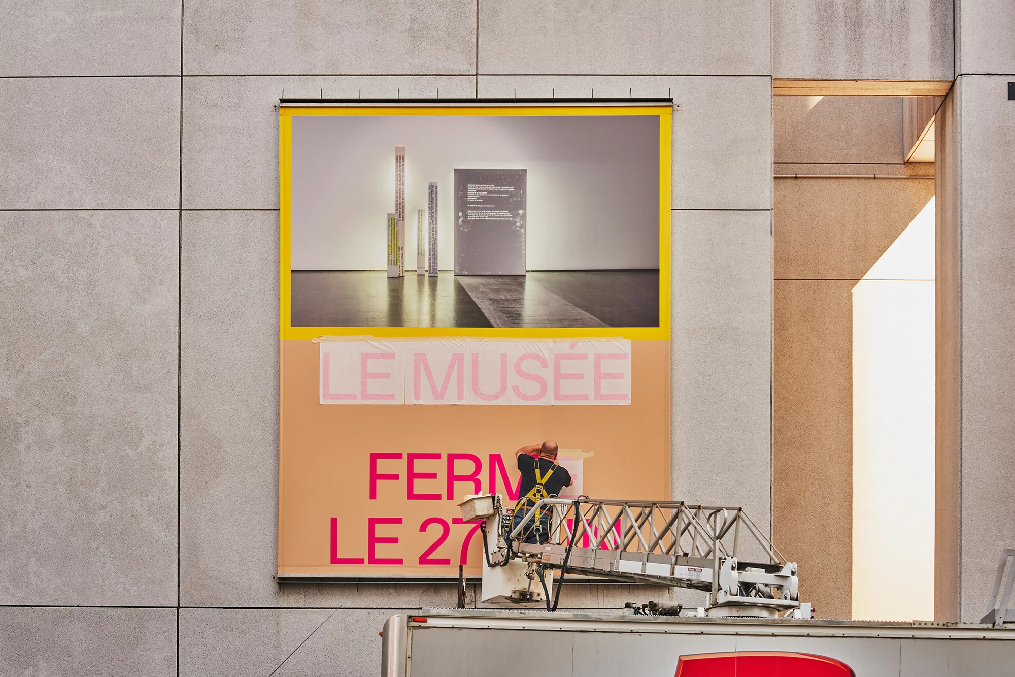 Adult male mounting museum poster with text 'LE MUSÉE FERMÉ LE 27' from a truck lift in urban setting, with concrete wall background on sunny day.