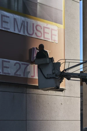 Person on a lift cleaning a large outdoor advertisement on the side of a building with pink lettering on a peach background.