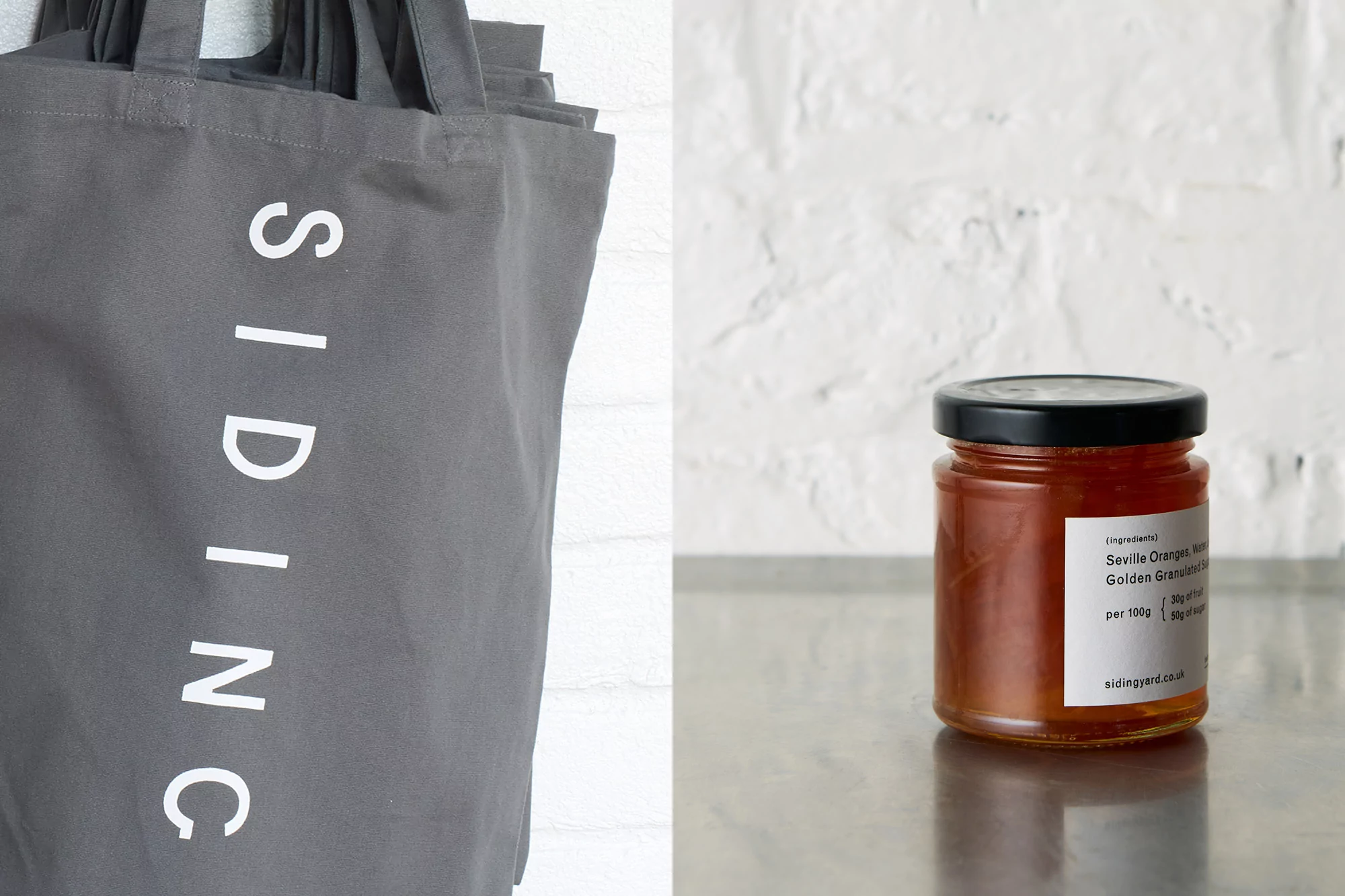 Promotional still life features a jar of Seville orange marmalade beside gray canvas tote bags displaying vertical white text logo.