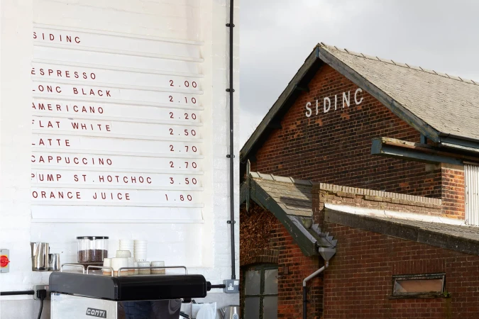 Coffee shop exterior featuring a menu board with drink prices next to a brick building with 'Siding' written on the side.