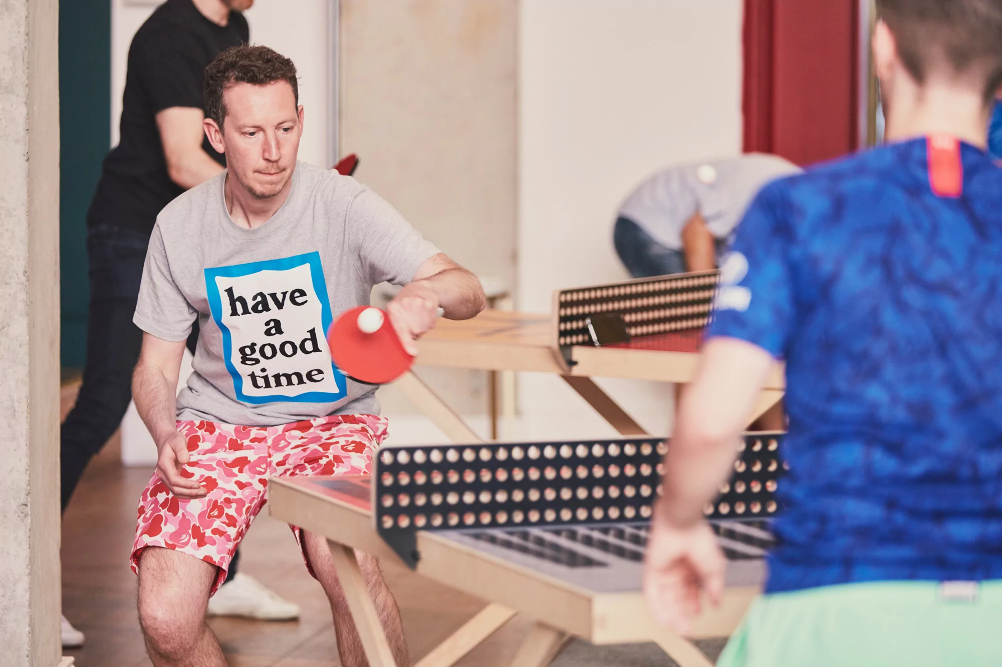 A man with a 'Have a Good Time' t-shirt plays ping pong with a red paddle, indoors, with other people in the background.