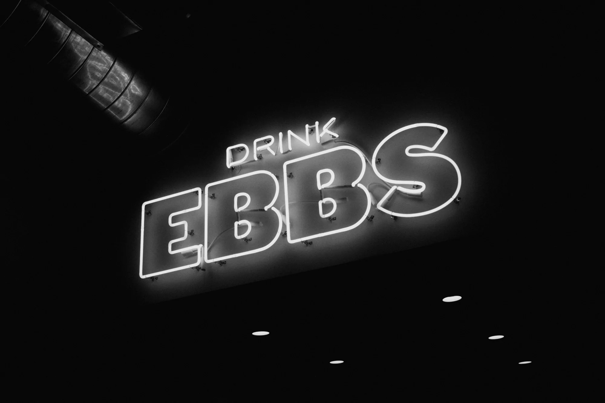 Monochrome shot of a neon sign that reads 'DRINK EBBS' against a black background, with industrial piping visible in the corner.
