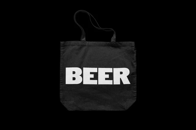 Black cotton tote bag with white “BEER” text graphic in a centered composition against a dark background with neutral lighting.