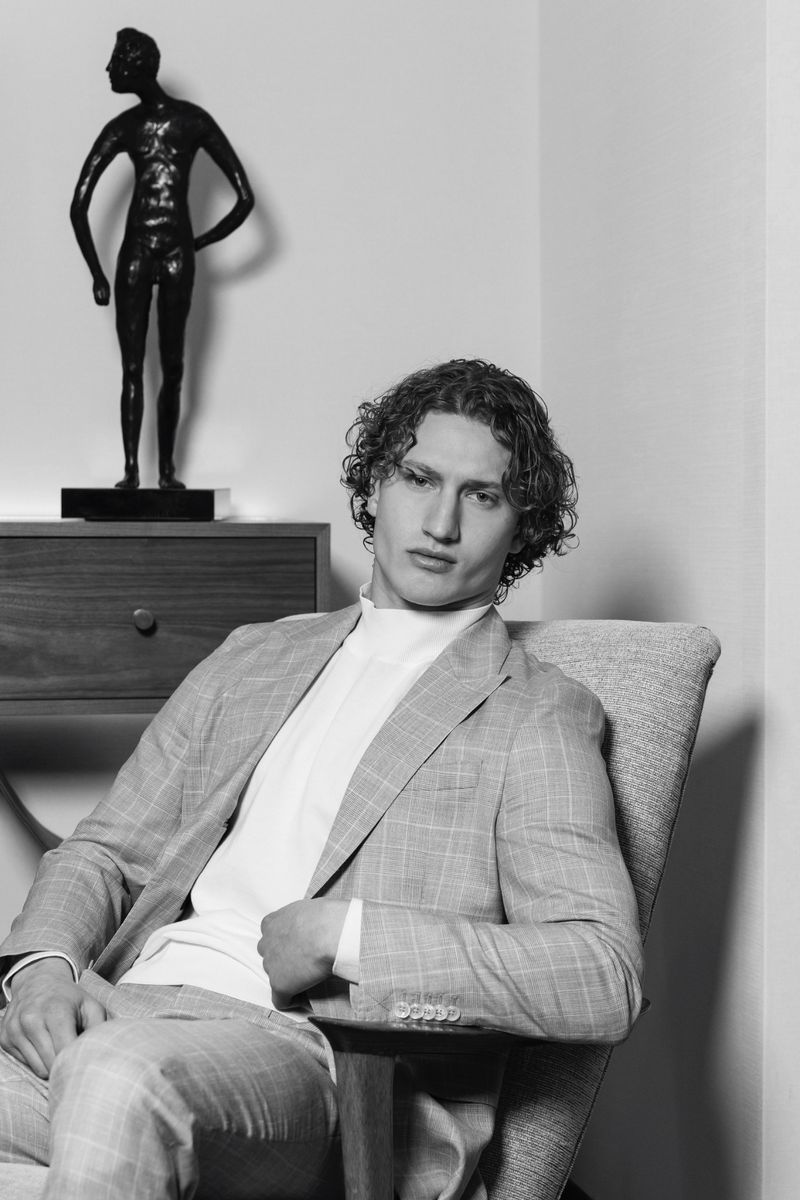 Monochrome portrait of a fair-skinned man in a grey plaid suit, seated in a chair with a sculpture and wooden cabinet behind.