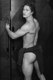 Black and white portrait of a male model posing by a dark paneled door with a sweater draped around his waist.