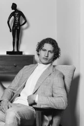 Portrait of a curly-haired male model seated in a chair wearing a plaid suit jacket for The Perfect Man Magazine.