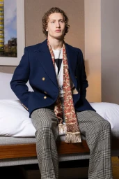 Model Brennan Aldred wearing a navy double-breasted blazer and patterned scarf seated indoors for The Perfect Man Magazine.