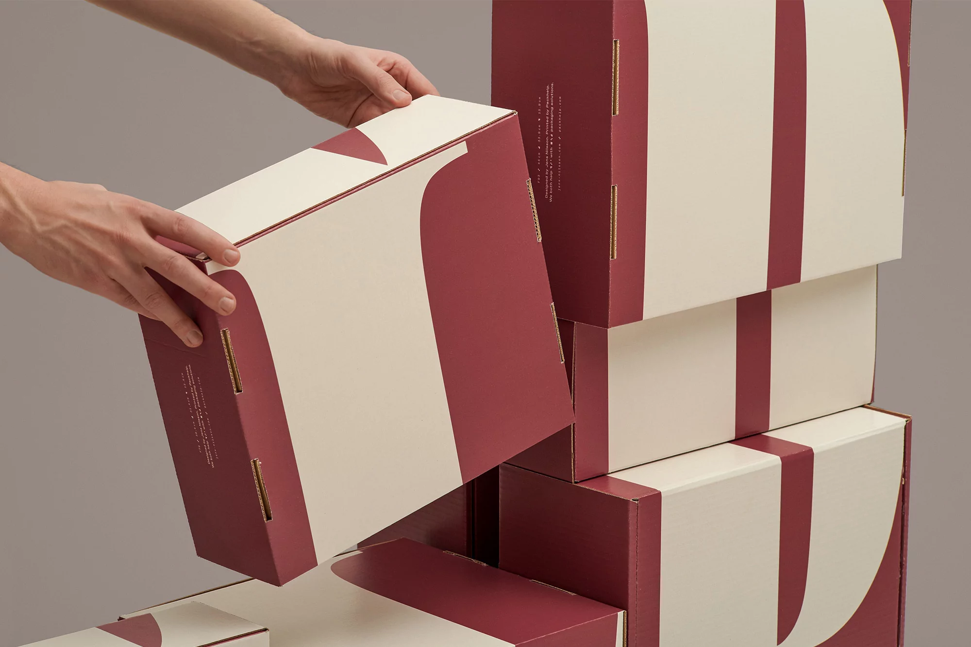 A stack of cream and burgundy boxes with graphic line design being held by a pair of hands in a studio with gray backdrop.