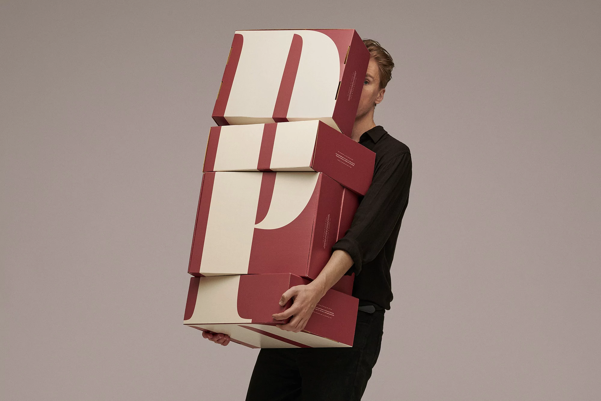 Adult man holding a stack of five red and cream-colored boxes with a letter 'D' printed on the front against a grey background.