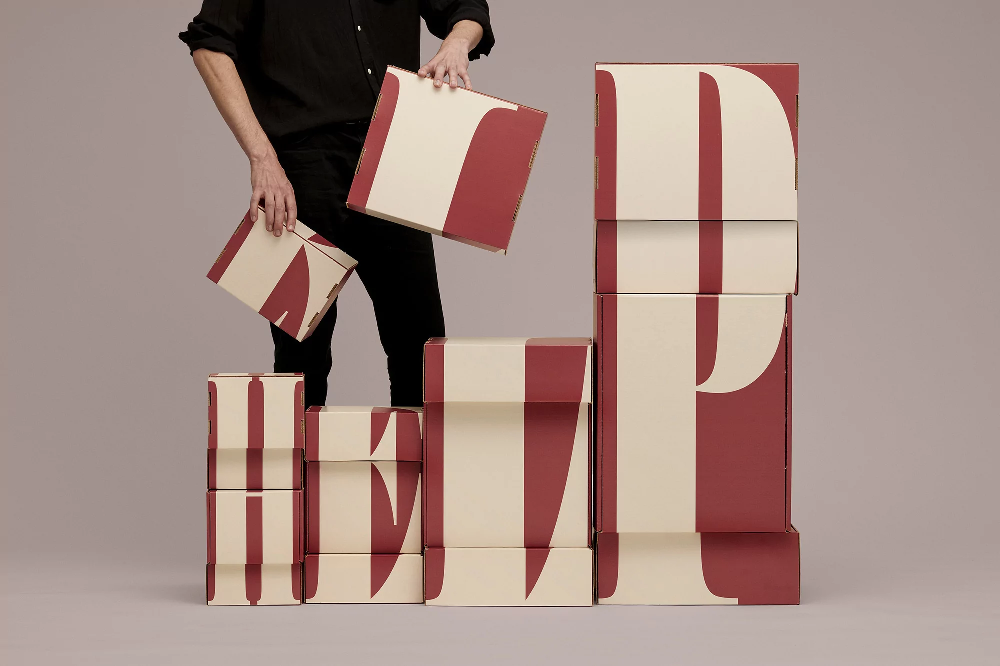 Person holding branded packaging boxes featuring bold typographic designs against a plain grey studio backdrop