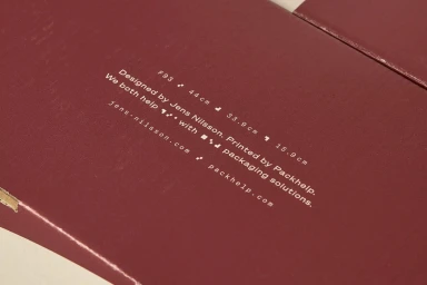 Close-up of a burgundy cardboard box surface featuring printed white typography and graphic details by Packhelp.
