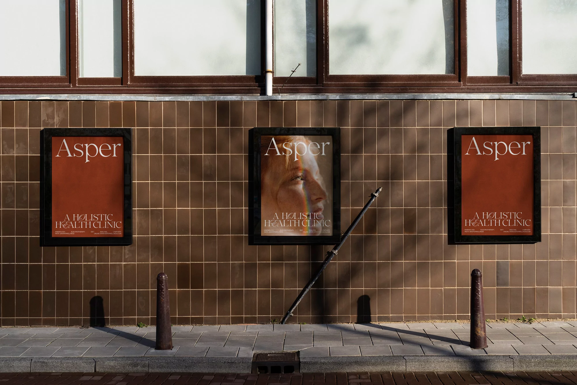 Three Asper holistic health clinic posters are mounted on a brown-tiled wall, with sunlight casting shadows on the sidewalk and wall.