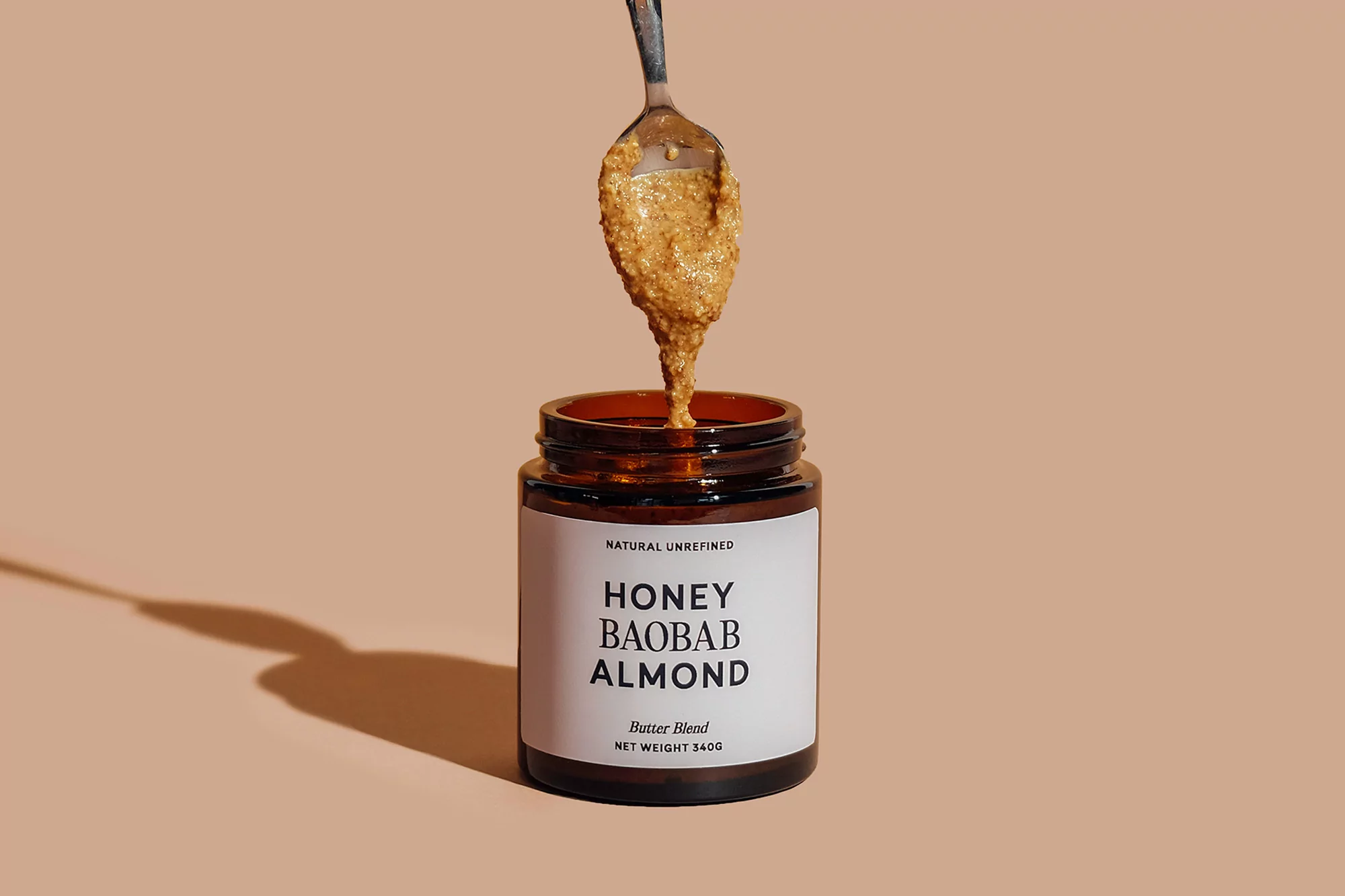 Amber jar of honey baobab almond butter with a spoonful dripping into the jar against a peach-colored background in a studio setting.