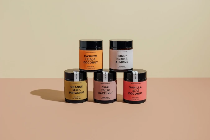 Six jars of nut butter stacked in a pyramid on a two-tone surface, featuring flavors like cashew chaga coconut, and vanilla acai coconut.