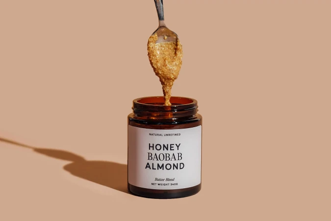 A spoon lifts textured nut butter above an amber glass jar labeled Honey Baobab Almond against a neutral beige background.
