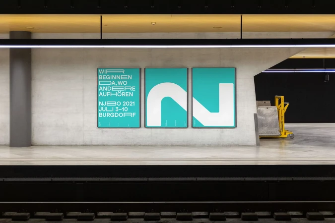 Three posters are mounted on a concrete wall at a train station platform, featuring white text and a graphic symbol on teal backgrounds.