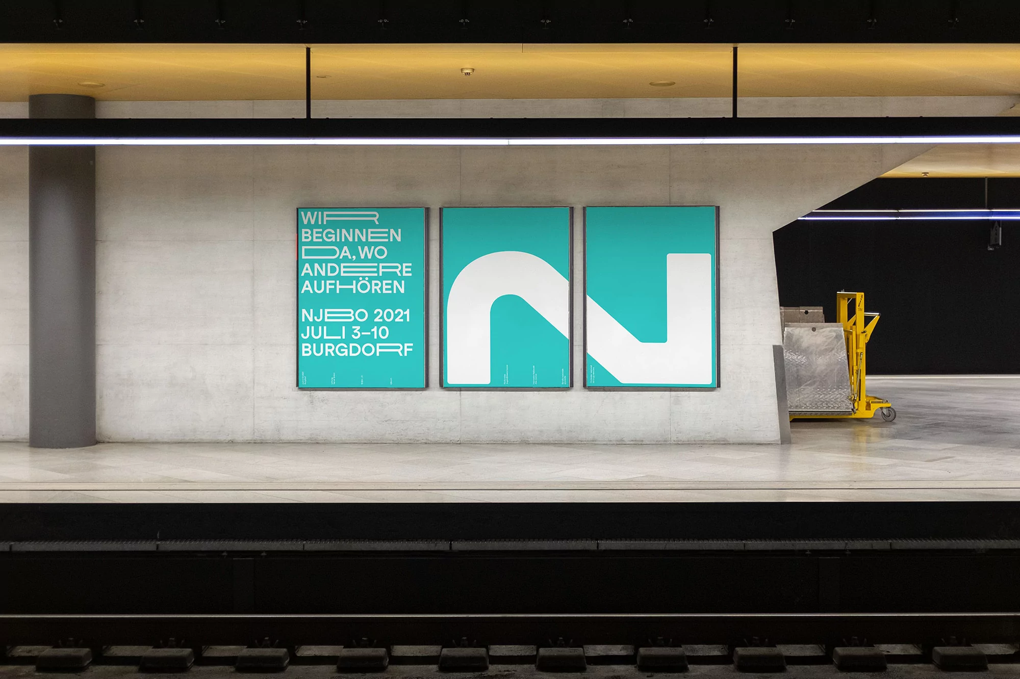 Three large teal and white posters mounted on a concrete wall in a modern underground transit station platform.
