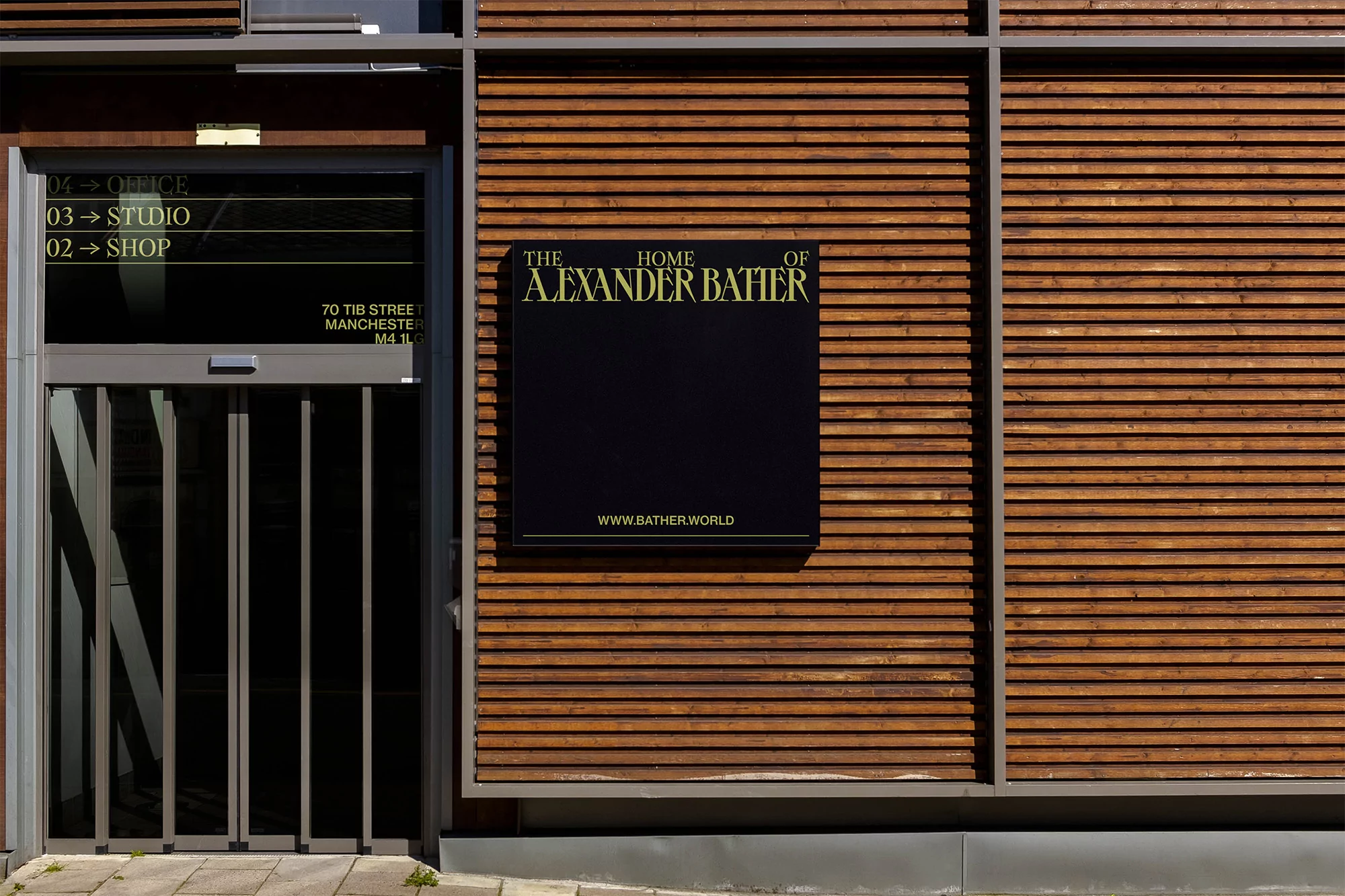 Exterior of a modern building featuring a dark sign for Alexander Bather, complemented by a gray door and a wooden-slatted facade.