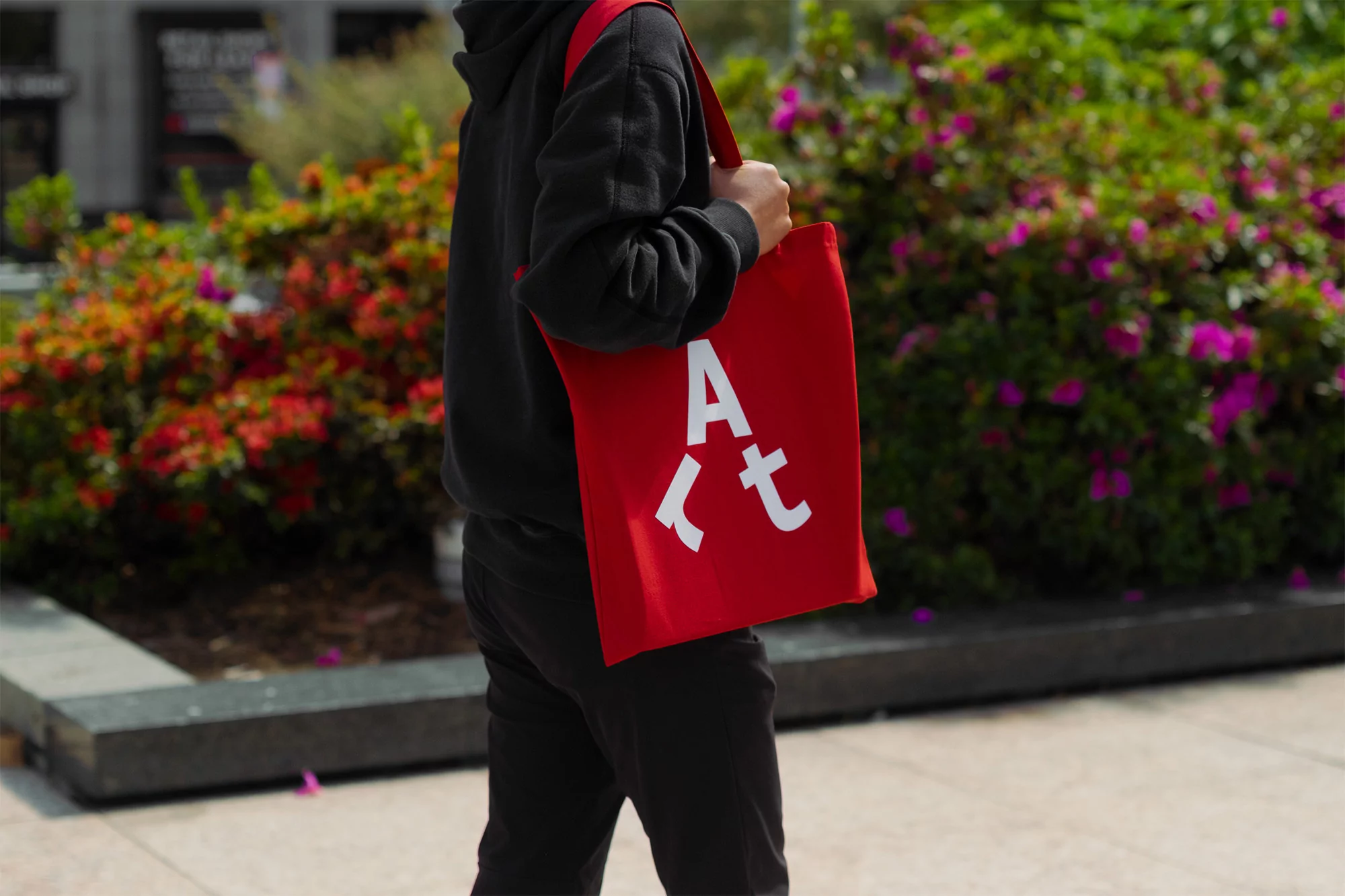 Person in black hoodie walks outdoors holding a red tote bag with white lettering, against a background of greenery and a grey sidewalk.