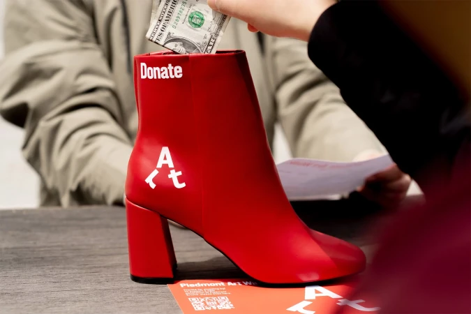 Red boot-shaped donation box with a hand inserting a dollar bill, promoting Piedmont Art Walk, captured in a medium shot.