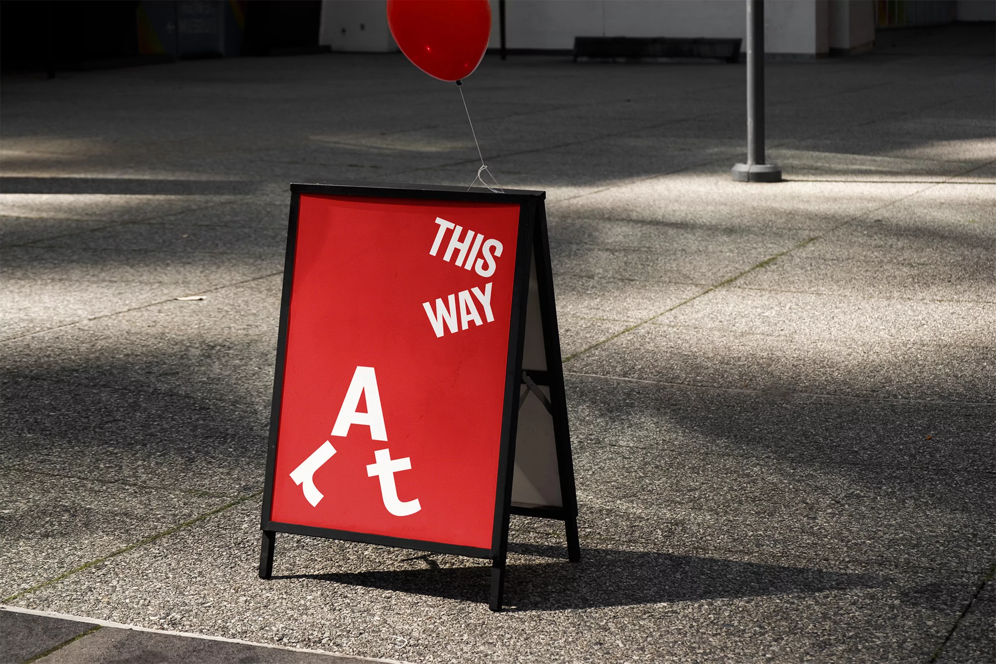 Red sign with white text reading 'This Way Art' on a concrete ground with a red balloon floating behind, bathed in sunlight.