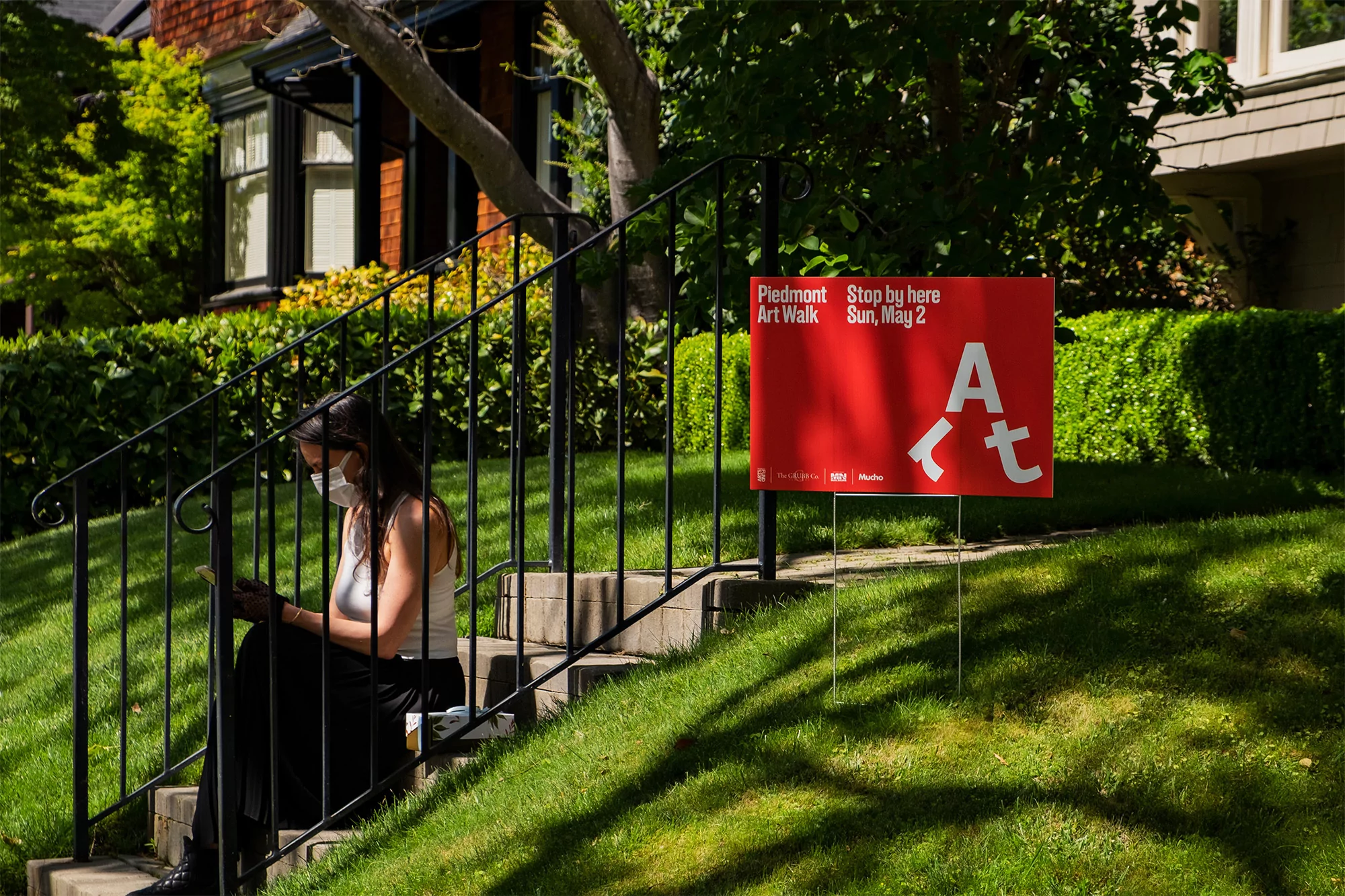 Woman sits on stairs with face mask, looking at phone, with a sign that reads 'Piedmont Art Walk' on a sunny day in residential area.