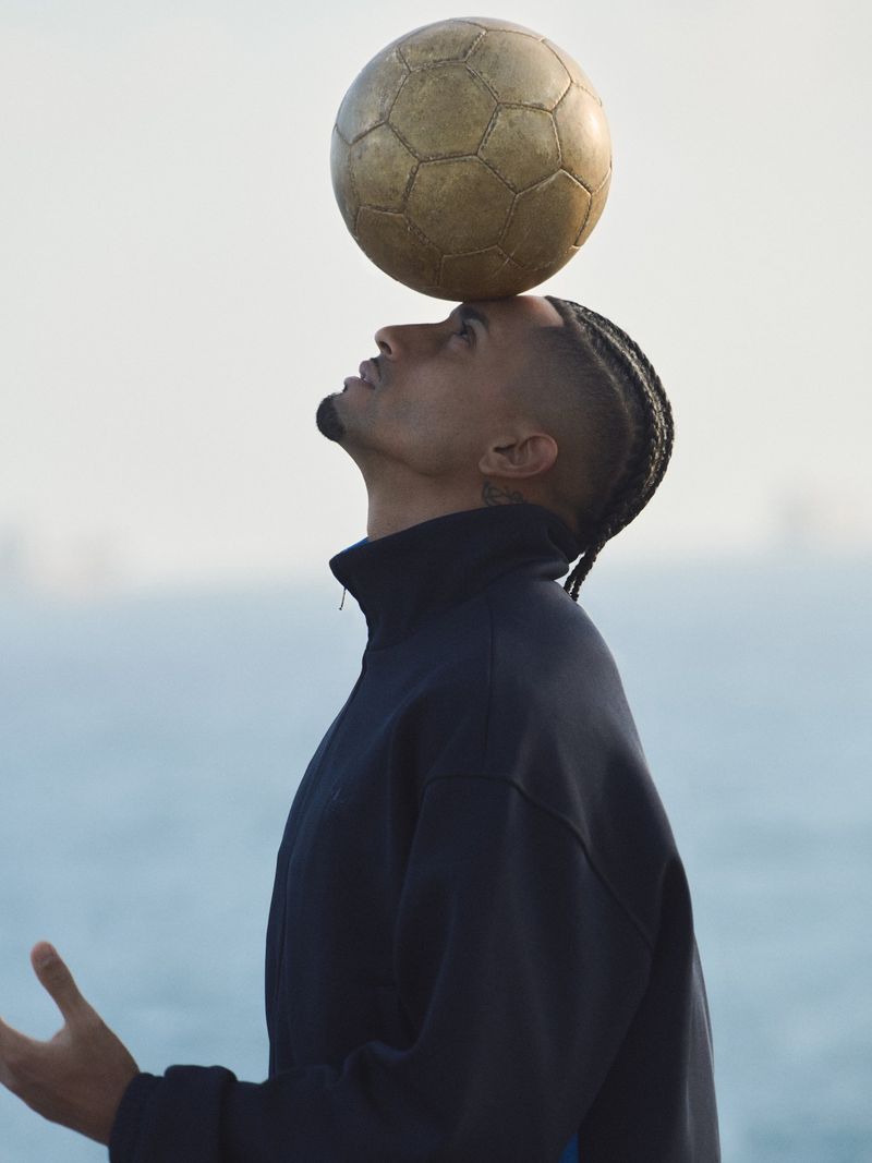 Adult male balances a soccer ball on his head outdoors near the ocean, wearing a blue jacket in a medium portrait.