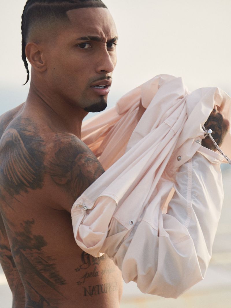 Adult man with tattoos poses shirtless outdoors, holding a light pink jacket in soft lighting. The background is neutral and blurred.