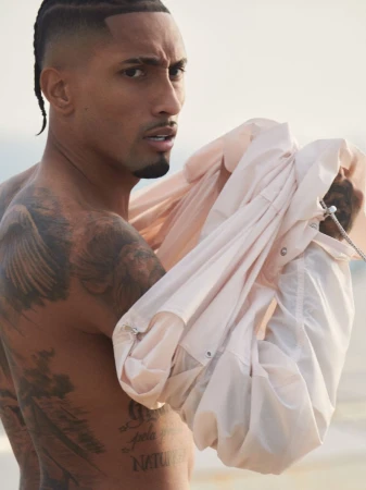 Adult man with tattoos poses shirtless outdoors, holding a light pink jacket in soft lighting. The background is neutral and blurred.