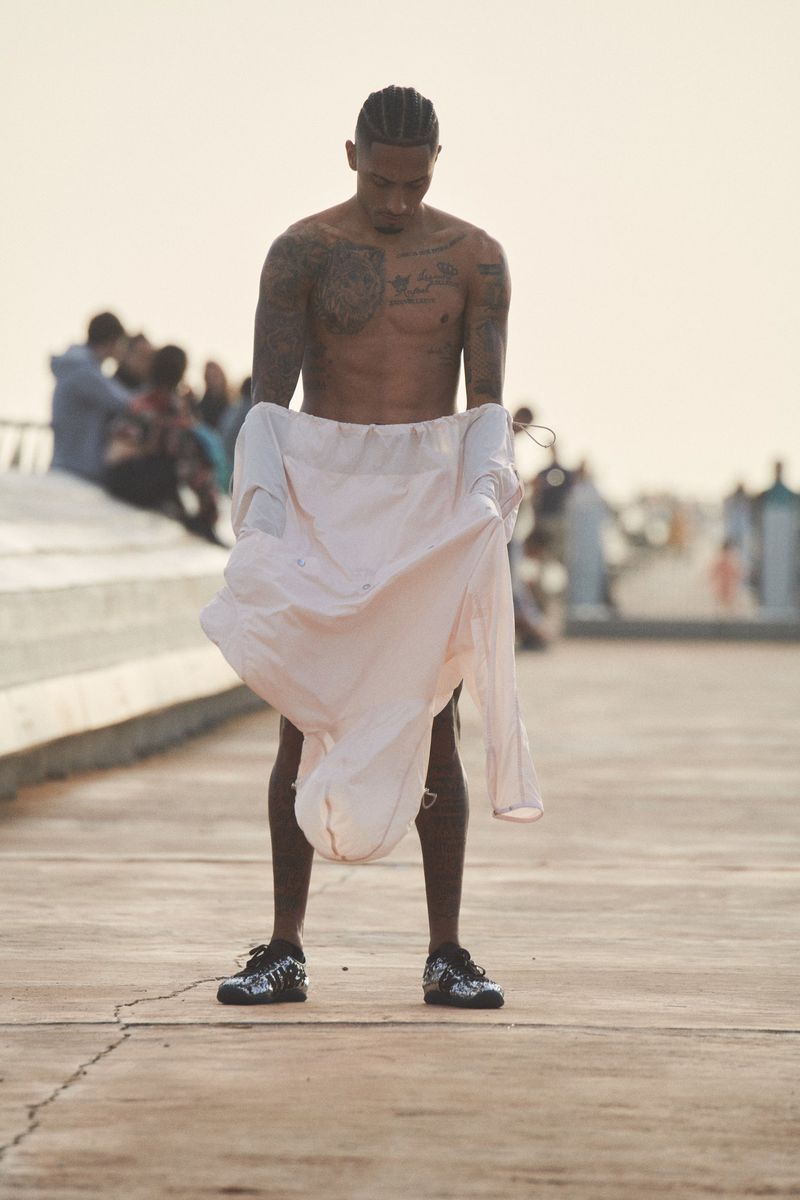 Tattooed man on boardwalk holding shirt outdoors. He wears shorts and sneakers with blurred background of people near the beach.