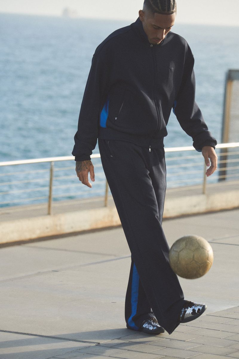 Adult male athlete in black sweatsuit balancing soccer ball outdoors on a sunny day near ocean, wearing shiny silver sneakers.
