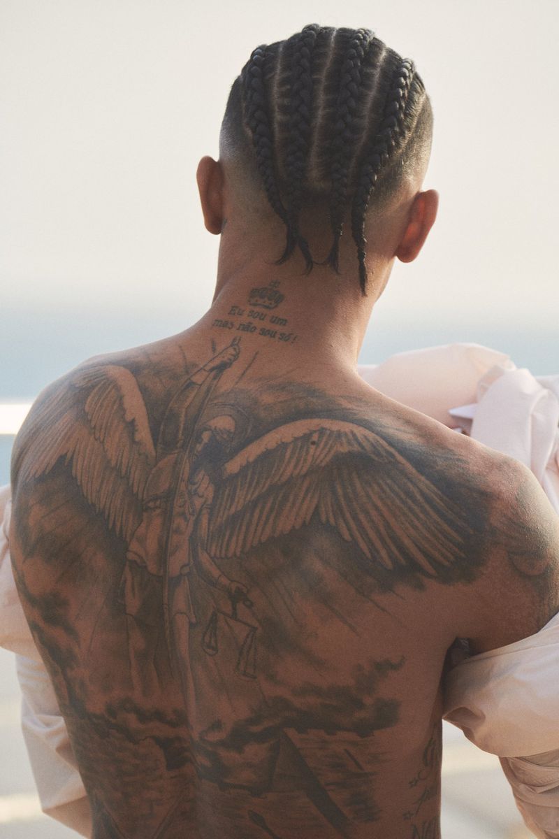 Man with braided hair and a detailed angel tattoo across his back, standing outdoors with ocean view, wearing a white shirt.