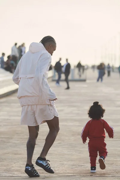 Man wearing a light pink sports jacket and shorts walking outdoors with a small child in a red tracksuit