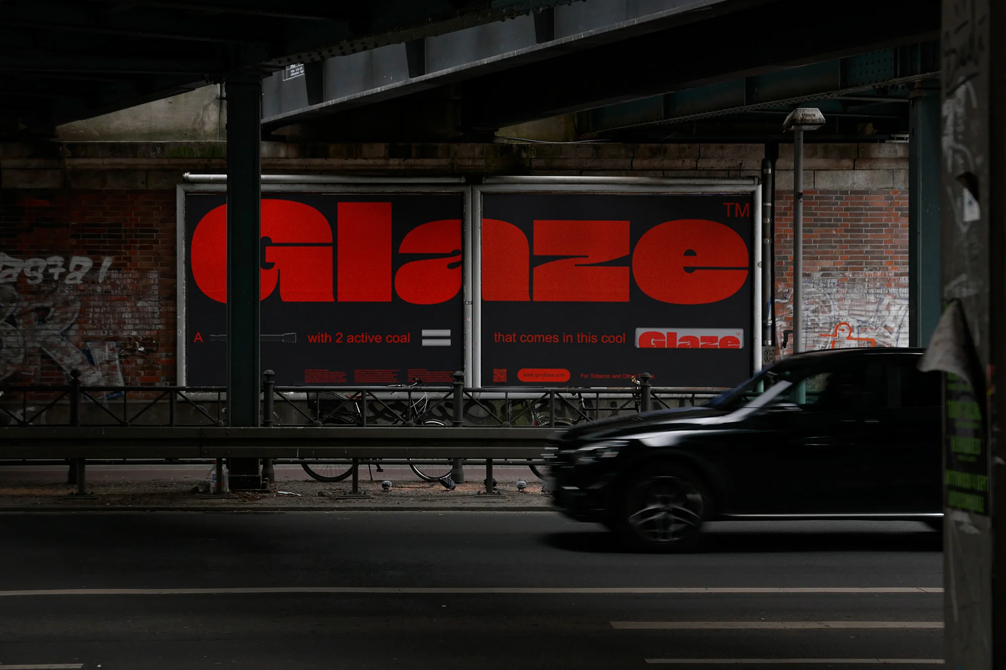 Black car drives along a dark urban street past a red billboard advertisement for Glaze, set against a brick building under a bridge.