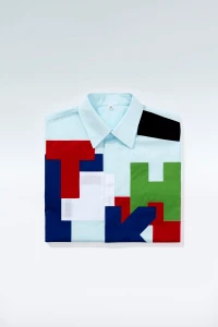 A folded light blue button-down shirt featuring colorful geometric letter blocks against a white studio background.