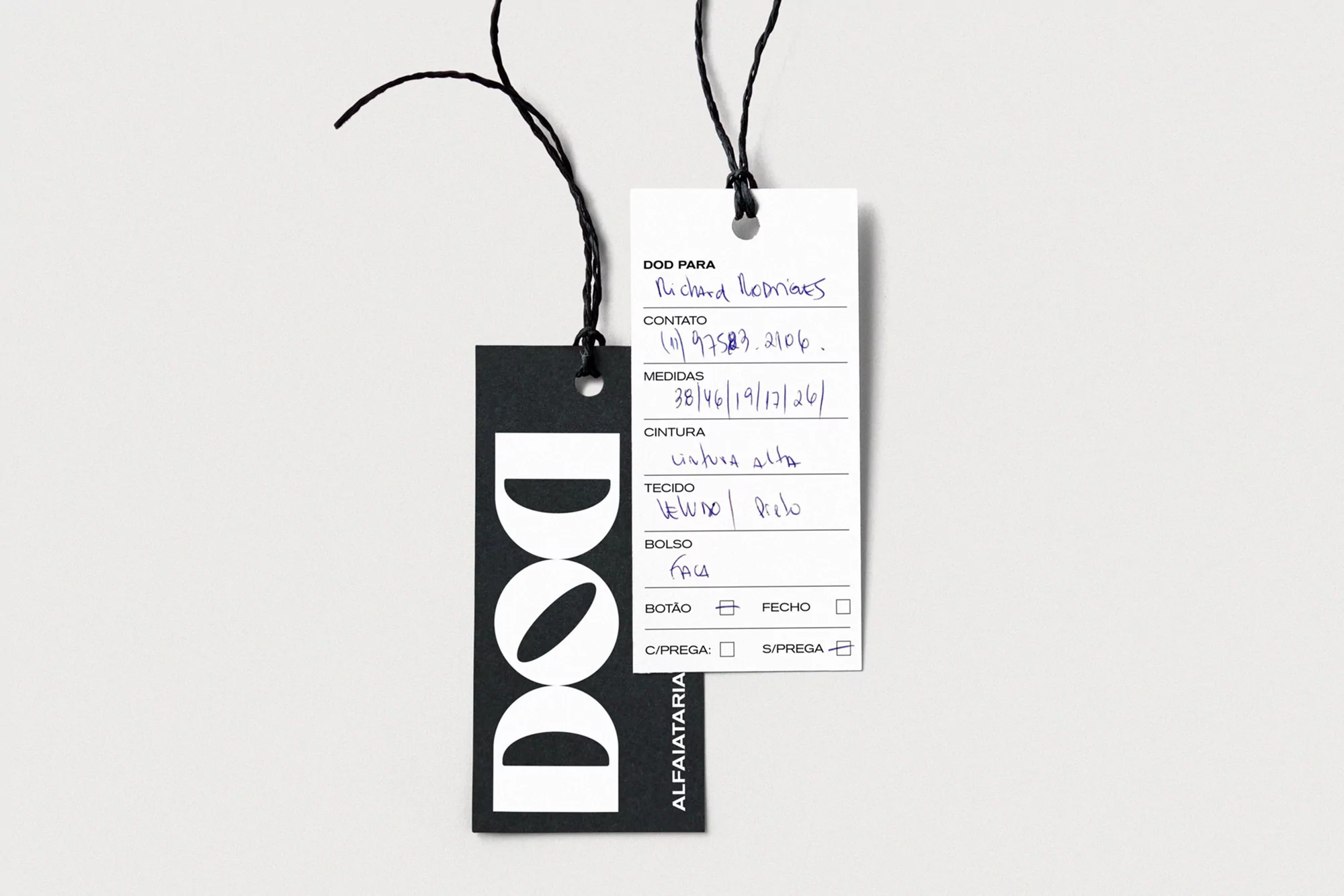Two clothing tags, one black with a white logo and one white with handwritten text, hanging from black strings against a white background.