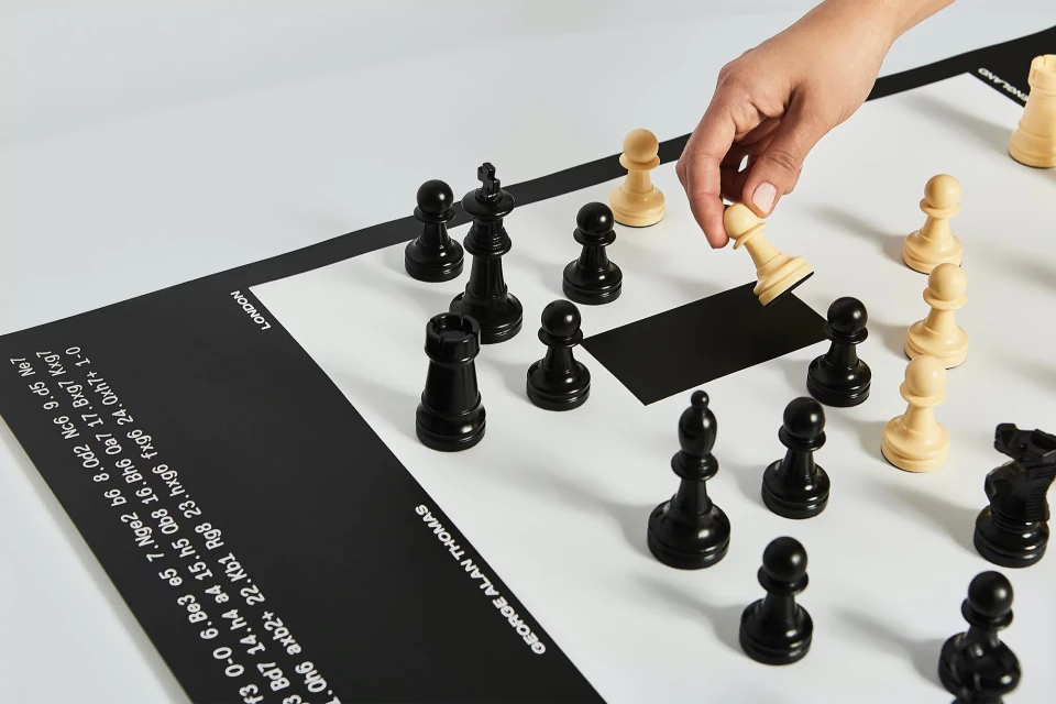Overhead shot of a person's hand moving a white pawn on a black-and-white chessboard with black chess pieces, featuring a modern design.