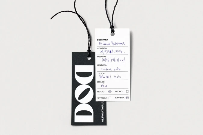Two paper clothing tags with black strings on a light grey background, featuring printed typography and handwritten blue ink.