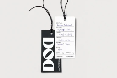 Two paper clothing tags with black strings on a light grey background, featuring printed typography and handwritten blue ink.