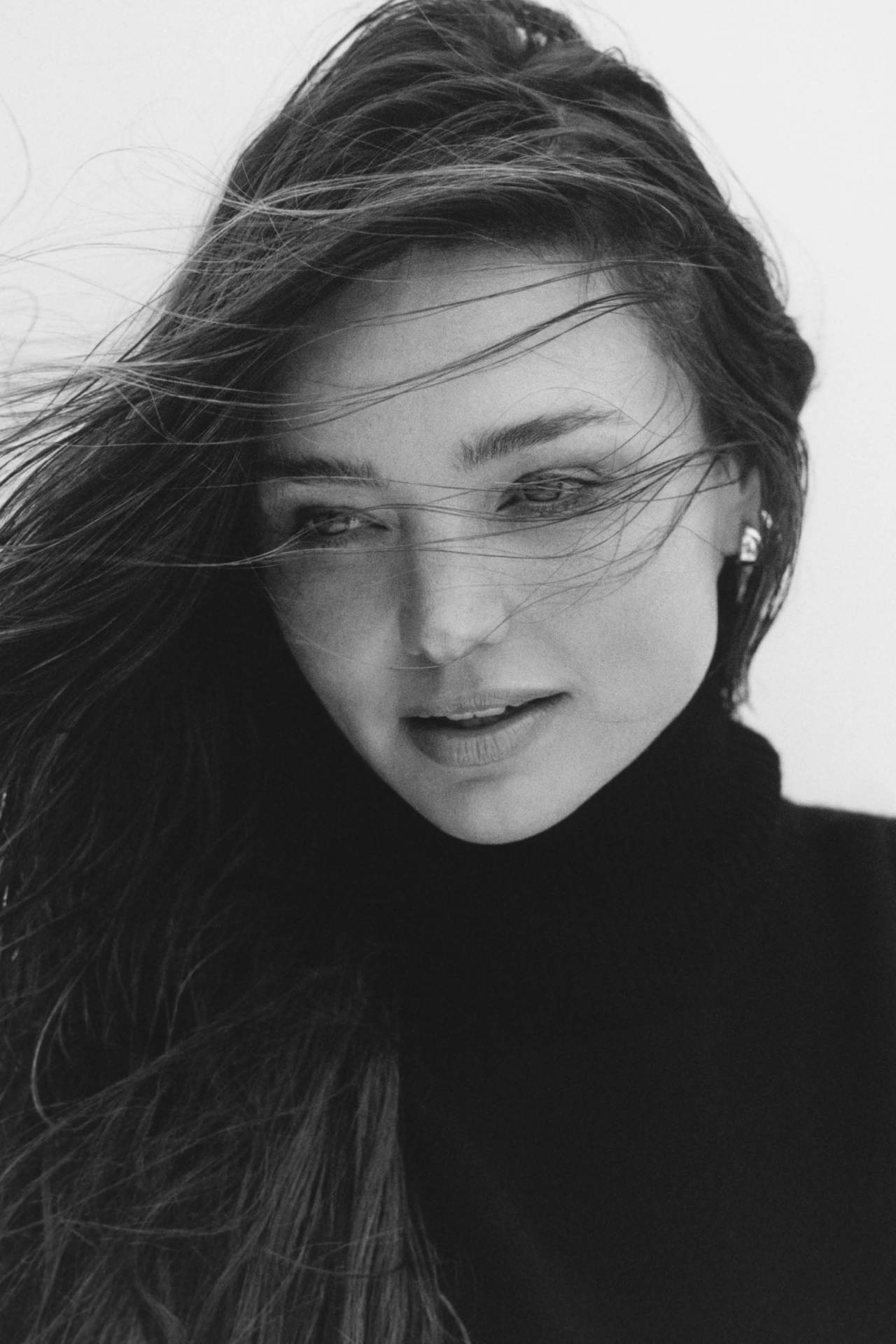Close-up portrait of Miranda Kerr with dark hair blowing across her face wearing a dark turtleneck against a plain background