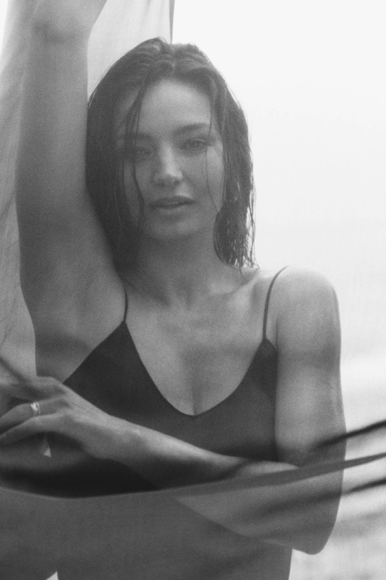 Monochrome portrait of an adult woman with wet hair in a slip dress, posing in front of a blurred background with soft, overcast lighting.