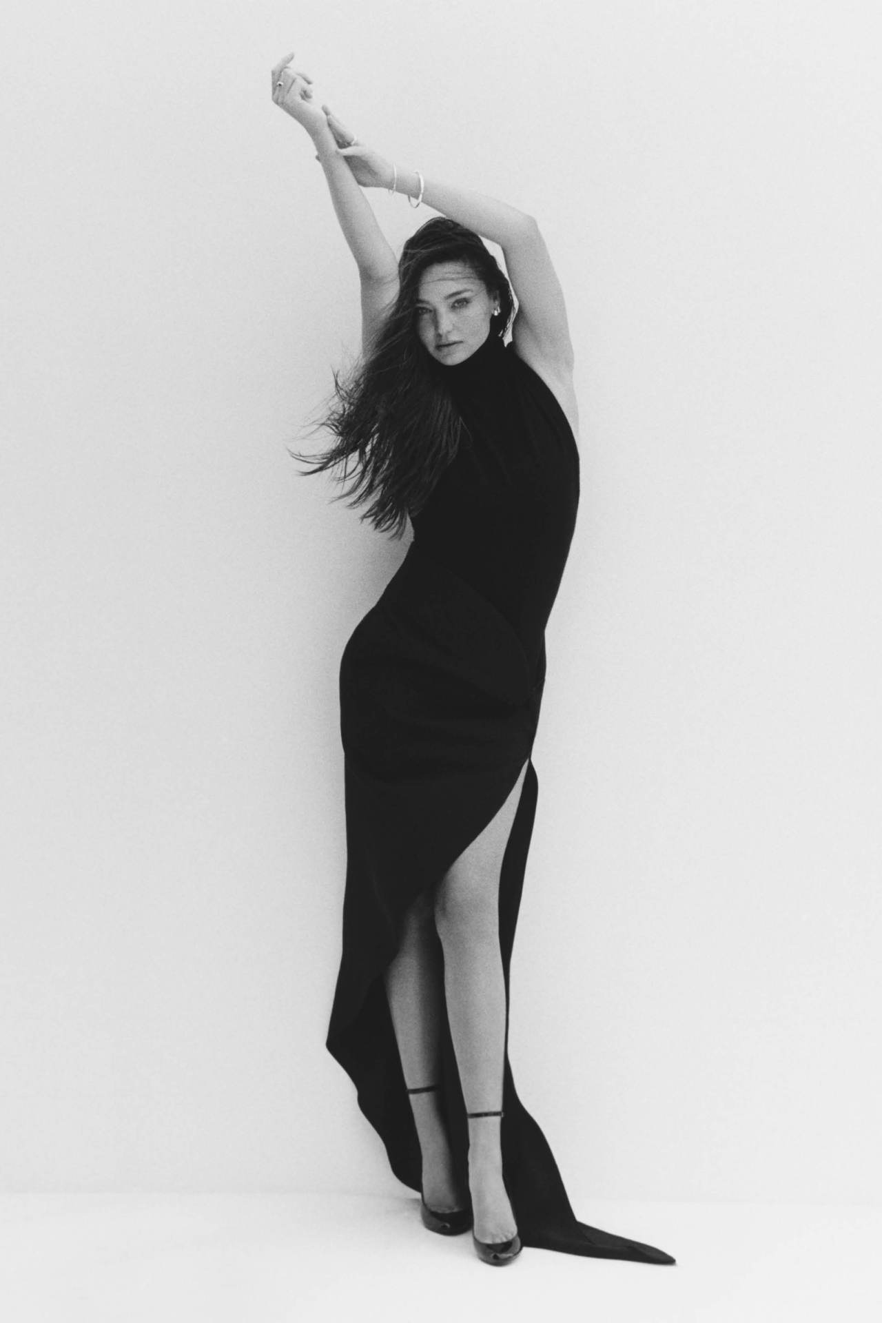 A woman poses in a black dress with a thigh-high slit against a white background, arms raised, showcasing an elegant and minimalist fashion style.