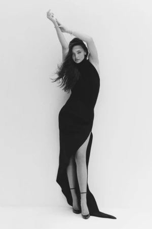 A woman poses in a black dress with a thigh-high slit against a white background, arms raised, showcasing an elegant and minimalist fashion style.