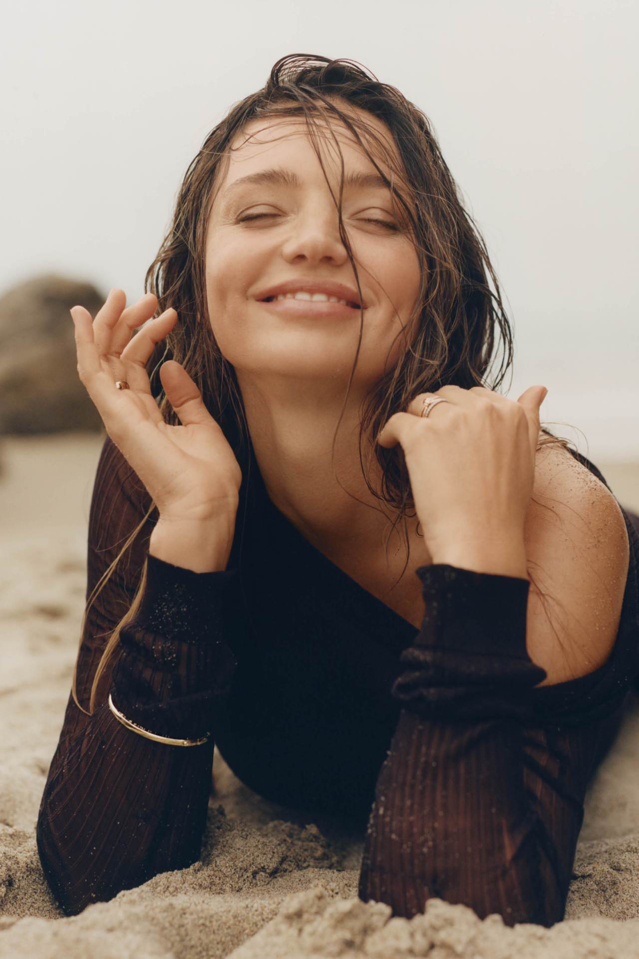 Smiling woman with wet hair lying on a sandy beach wearing a dark long-sleeve top
