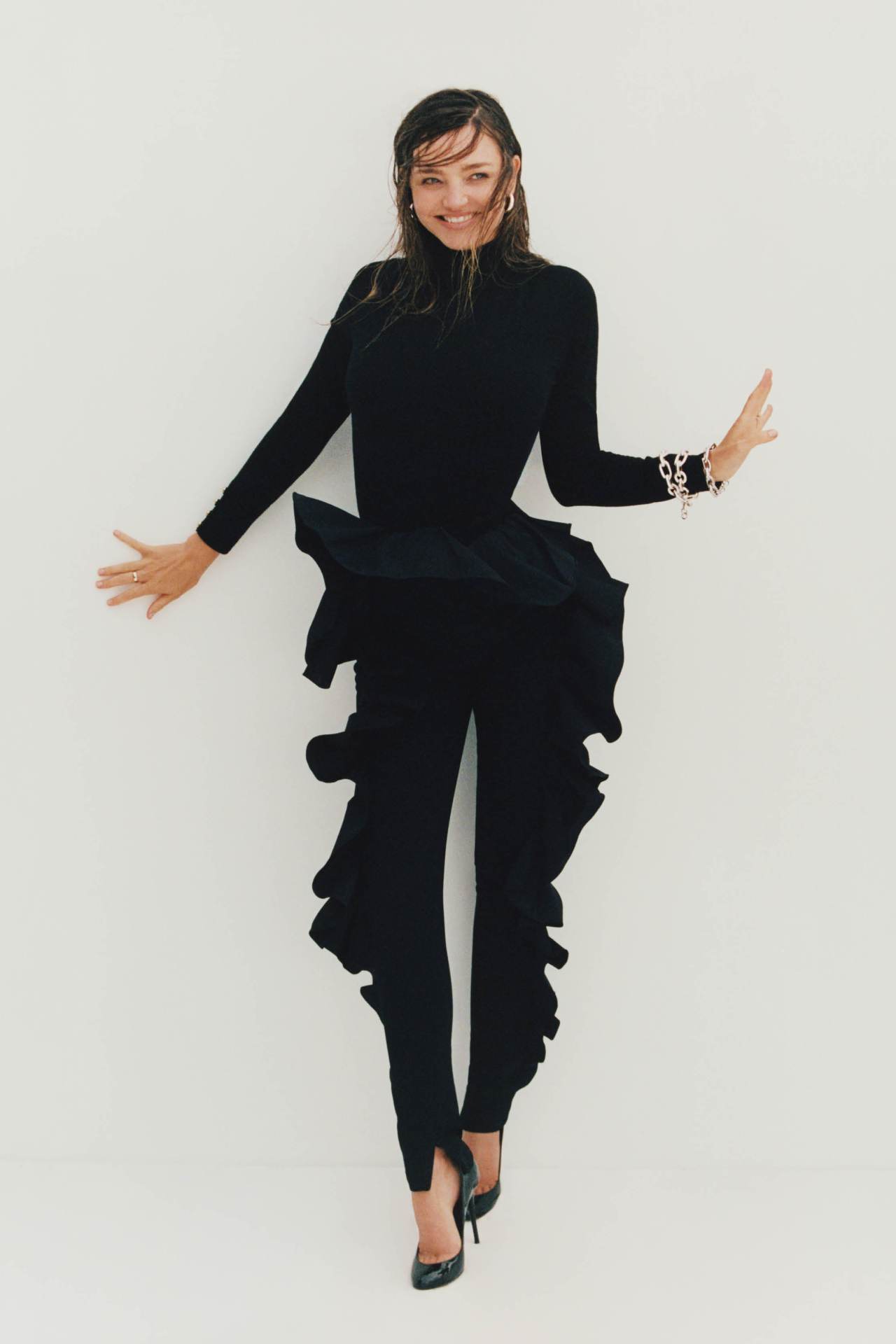 A woman in a long-sleeved black top and ruffled black pants poses against a white background with an open-arm gesture and a smile.