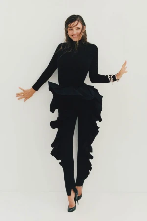 A woman in a long-sleeved black top and ruffled black pants poses against a white background with an open-arm gesture and a smile.