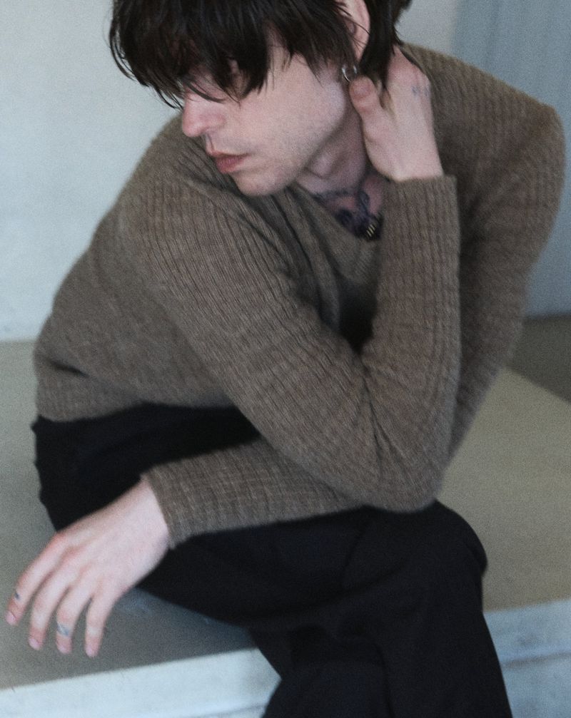 Young man in a brown ribbed sweater and black pants sits on concrete steps in a fashion portrait. The man is fair-skinned with dark hair.