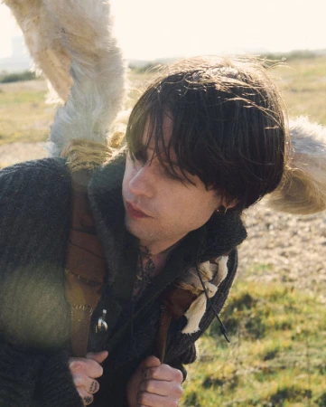 Adult male with dark hair poses outdoors wearing a gray coat and unique faux fur shoulder accessory in a natural, outdoor setting.