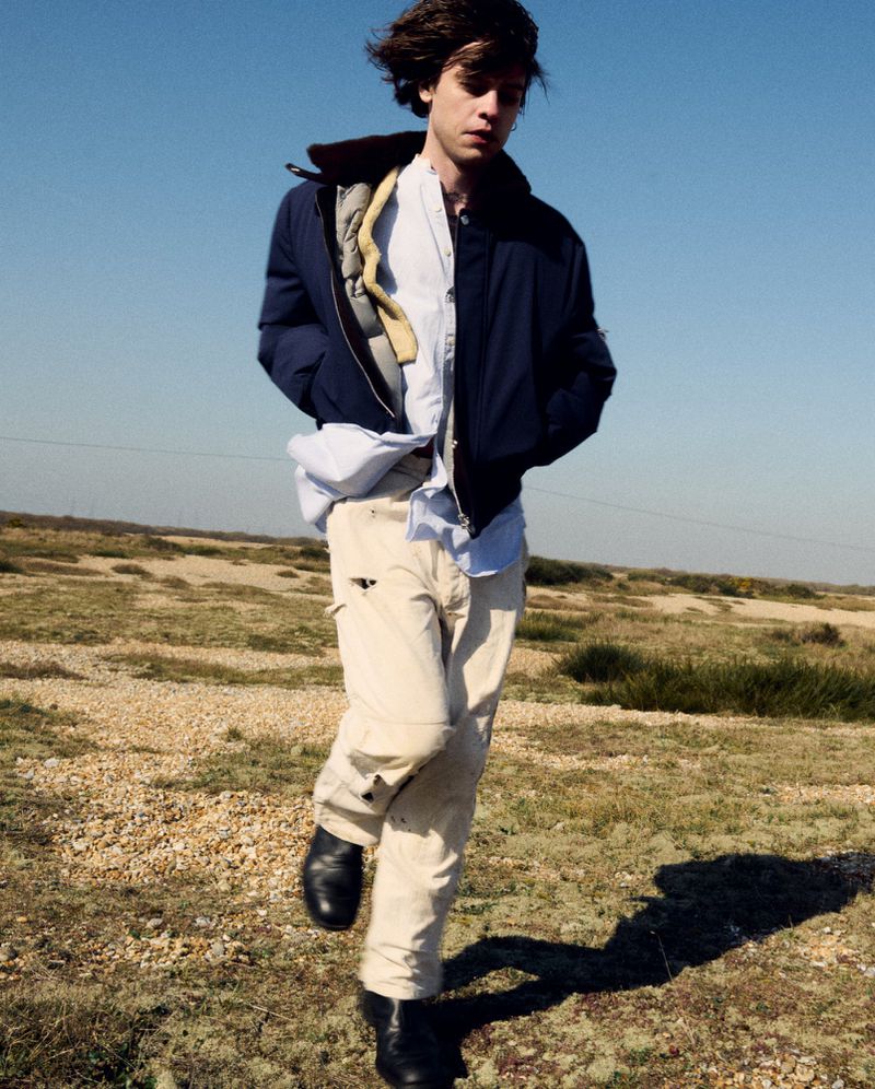 A young adult male strides confidently across a dry, grassy landscape beneath a clear blue sky, showcasing casual fashion in an outdoor setting.