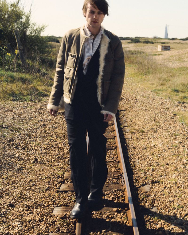 An adult male walks along a railroad track wearing a tan jacket, dark pants, and black shoes in a rural setting during daylight hours.
